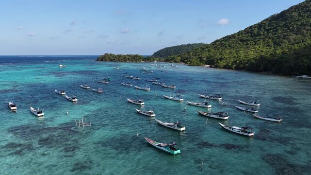 Aerial view of numerous small fishing boats anchored in the clear turquoise waters of a bay, surrounded by lush green hills in Karimunjawa, Central Java, Indonesia.