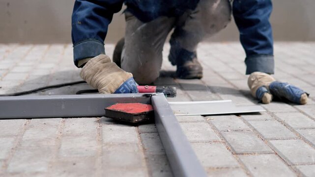 Low-angle view shows gloved worker kneeling over gray metal support on light brick paving, using handheld cutting tool with bright contact glow, faint smoke, and practical maintenance mood