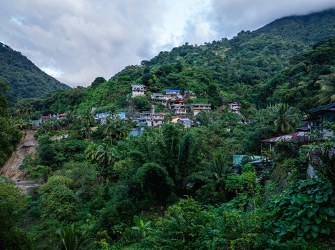 Aerial view of a mountain village nestled in lush green hills with dense vegetation, scattered houses, and steep terrain under a cloudy sky in Tuba, Cordillera Administrative Region, Philippines.