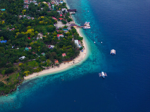 Aerial view of the coastline featuring a sandy beach, lush vegetation, and traditional outrigger boats floating in clear turquoise waters in Santander, Central Visayas, Philippines.
