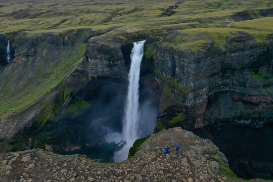 Aerial view of the majestic Haifoss waterfall plunging into a deep canyon with people standing on the rocky cliff edge in the foreground in Hella, Rangarthing ytra, Iceland.