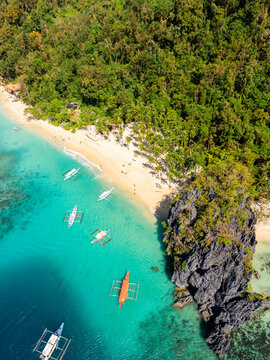 Aerial view of Seven Commandos Beach with turquoise waters, white sand, lush tropical forest, rocky cliffs, and traditional outrigger boats in El Nido, Palawan, Philippines.