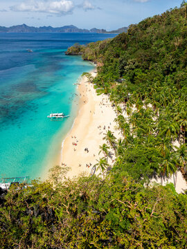 Aerial view of Seven Commandos Beach with turquoise water, white sand, lush palm trees, and traditional outrigger boats in El Nido, Palawan, Philippines.