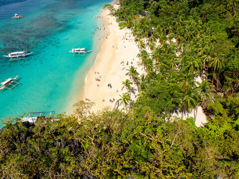 Aerial view of Seven Commandos Beach with white sand, turquoise water, traditional outrigger boats, and lush palm trees in El Nido, Palawan, Philippines.