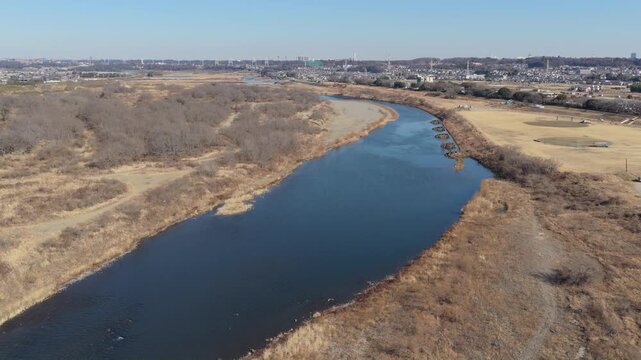 Aerial view of a winding river flowing through a dry landscape with sparse trees and distant suburban buildings under a clear blue sky in Japan.
