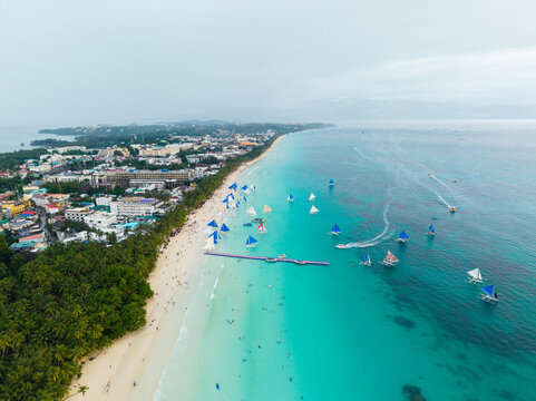 Aerial view of Boracay island featuring white sand beaches, turquoise water, numerous sailboats, and lush palm trees along the coastline in Malay, Western Visayas, Philippines.