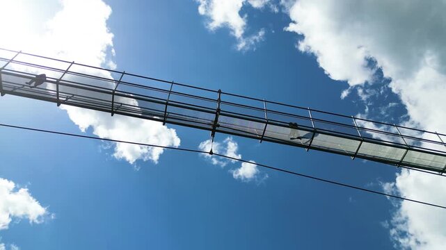 Aerial view of the Sky Bridge 721 suspension bridge with people walking across it against a bright blue sky with clouds in Dolni Morava, Pardubicky kraj, Czechia.