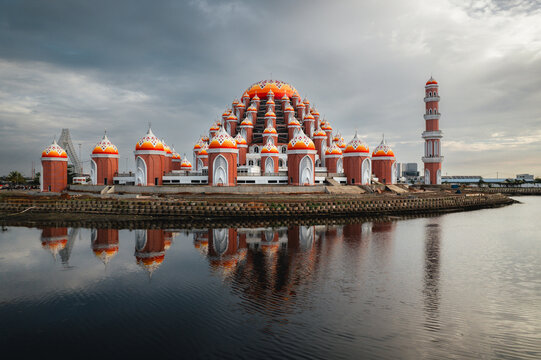 Aerial view of Masjid Kubah 99 Asmaul Husna with its distinctive orange domes and minaret reflecting in the calm water in Kecamatan Mariso, Sulawesi Selatan, Indonesia.