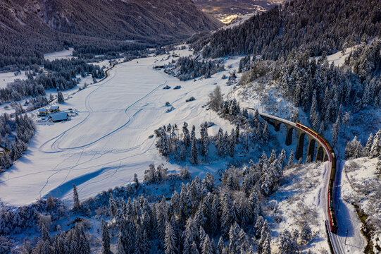 Aerial view of the Glacier Express train crossing the Landwasser Viaduct through a snow-covered valley surrounded by pine forests in Bergun Filisur, Graubunden, Switzerland.
