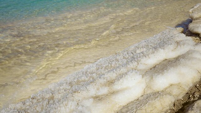 Crystalline salt covering completely the sandy shore of Dead Sea on Jordan side, closeup detail to sharp hard crystals, clear water small waves near