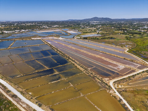 Aerial view of the salt pans in Moncarapacho with geometric pools of varying water colors and dirt paths winding through the landscape in Moncarapacho, Faro District, Portugal.