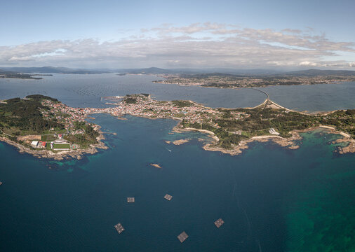 Aerial view of A Illa de Arousa surrounded by the blue waters of the Atlantic Ocean, featuring the connecting bridge, coastal town, and mussel rafts in A Illa de Arousa, Pontevedra, Spain.
