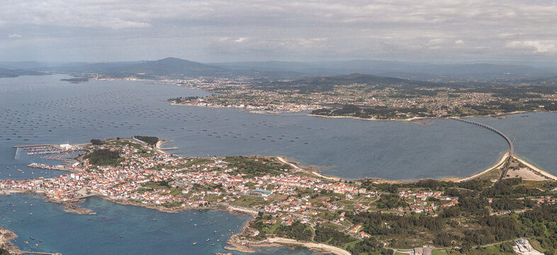 Aerial view of the town of A Illa de Arousa with red-tiled roofs, connected by a bridge to the mainland and surrounded by mussel rafts, A Illa de Arousa, Pontevedra, Spain.