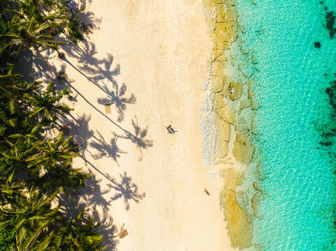 Aerial view of Puka Shell Beach in Boracay with palm trees casting long shadows on white sand, turquoise water, and rocky shoreline with people walking in Malay, Western Visayas, Philippines.