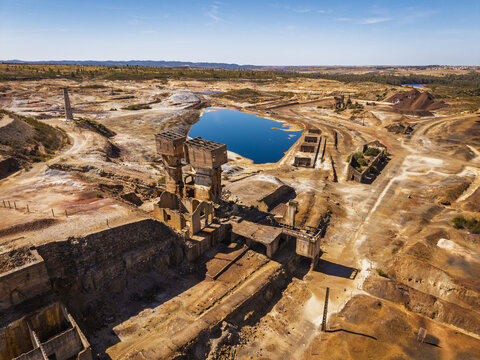 Aerial view of the abandoned Achada do Gamo mine site featuring industrial ruins, a vibrant blue water-filled pit, and arid terrain in Achada do Gamo, Beja, Portugal.