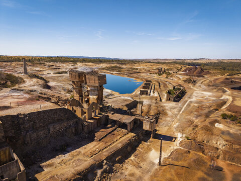 Aerial view of the abandoned Achada do Gamo mine site featuring industrial ruins and a vibrant blue water-filled pit set within an arid landscape in Achada do Gamo, Beja, Portugal.