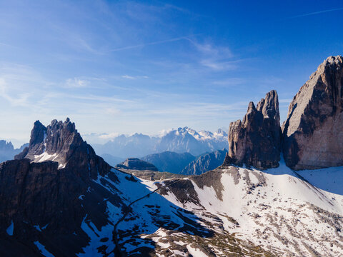 Aerial view of the Tre Cime di Lavaredo peaks with rocky slopes, patches of snow, and a clear blue sky in the Dolomites, Italy.