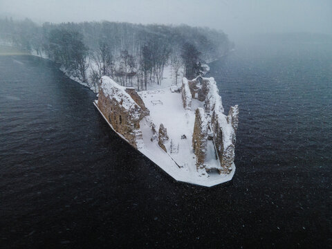 Aerial view of Koknese Castle Ruins covered in snow during a blizzard with surrounding water and forest in Koknese, Aizkraukles novads, Latvia.