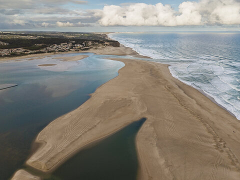 Aerial view of the Foz do Arelho lagoon meeting the Atlantic Ocean with sandy beaches, calm turquoise waters, and rolling waves along the coastline in Foz do Arelho, Leiria District, Portugal.