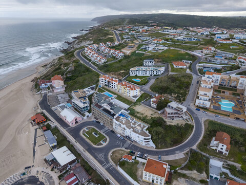 Aerial view of the coastal town of Foz do Arelho with its sandy beach, modern apartment buildings, winding roads, and surrounding green hills in Foz do Arelho, Leiria District, Portugal.