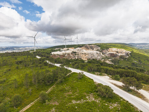 Aerial view of Parque Eolico da Serra dos Candeeiros featuring wind turbines and an open-pit limestone quarry surrounded by green hills and a winding road in Rio Maior, Santarem, Portugal.