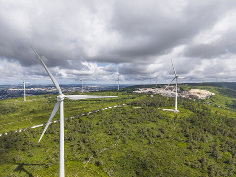 Aerial view of the wind turbines at Parque Eolico da Serra dos Candeeiros, set amidst lush green hills and a cloudy sky in Rio Maior, Santarem, Portugal.