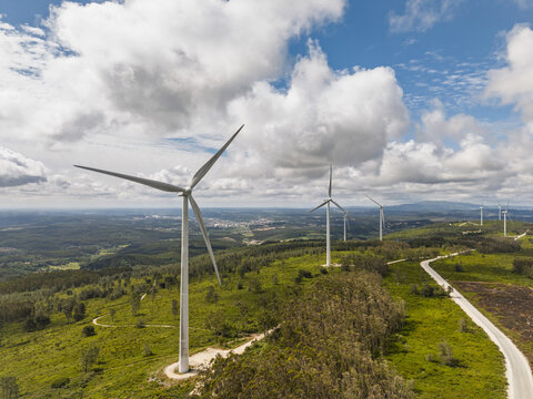 Aerial view of wind turbines at Parque Eolico da Serra dos Candeeiros, set against green hills, winding roads, and a blue sky with clouds, Rio Maior, Santarem, Portugal.