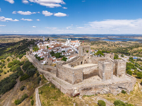Aerial view of Monsaraz Castle and the historic village with white-washed houses, stone walls, and surrounding landscape under a bright blue sky in Monsaraz, Evora District, Portugal.