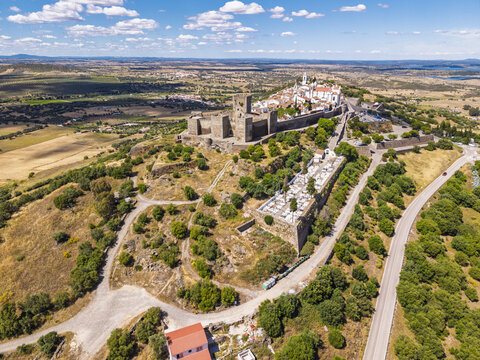 Aerial view of the historic Monsaraz castle and village perched on a hilltop, surrounded by rolling countryside, olive groves, and a cemetery in Monsaraz, Evora District, Portugal.