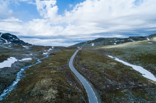 Aerial view of the winding Aurlandsfjellet road cutting through a rugged mountainous landscape with patches of snow, a flowing stream, and a dramatic cloudy sky in Aurland, Vestland, Norway.