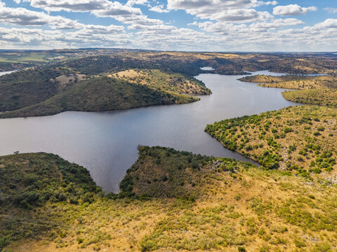 Aerial view of Albufeira do Alqueva reservoir with rolling hills and dry vegetation under a blue sky in Alqueva, Distrito de Evora, Portugal.