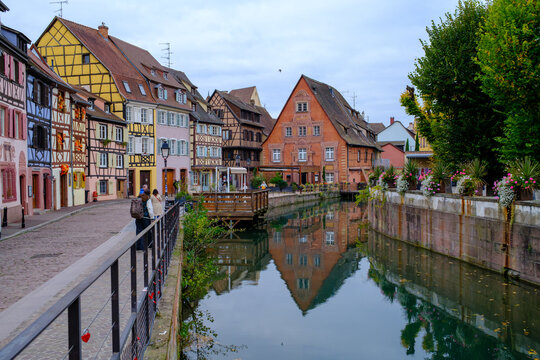 Colmar, France - 09 July 2026: View of the historic half-timbered houses along the canal in La Petite Venise with people walking on the cobblestone street and reflections in the water.
