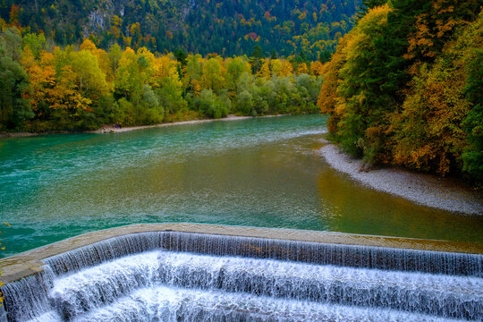View of the cascading water of a weir on the Lech river surrounded by vibrant autumn foliage and dense forest in Fussen, Bavaria, Germany.