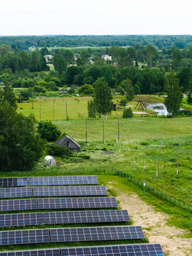Aerial view of a solar farm with rows of photovoltaic panels set in a lush green rural landscape with trees, a small pond, and a wooden structure in Suntazi, Ogre Municipality, Latvia.