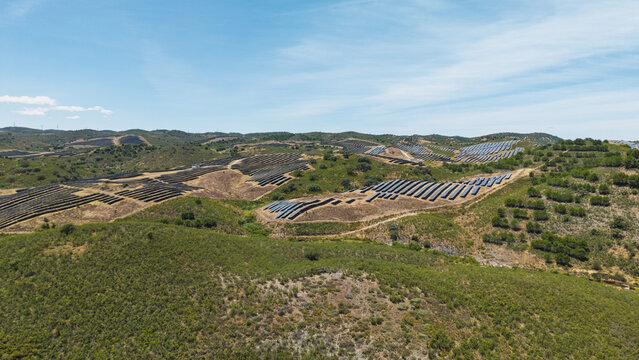 Aerial view of a large solar farm with rows of photovoltaic panels installed on rolling green hills under a clear blue sky in Vaqueiros, Faro District, Portugal.