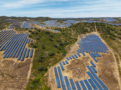 Aerial view of a large solar farm with rows of photovoltaic panels installed across the rolling hills of the countryside under a clear blue sky in Vaqueiros, Faro District, Portugal.