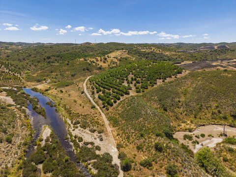Aerial view of the rural landscape near Vaqueiros, featuring a winding river, rolling hills, and orchards under a clear blue sky in Vaqueiros, Faro District, Portugal.