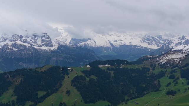 Aerial view of Stanserhorn Cabrio with snow-capped mountain peaks and lush green alpine slopes under a cloudy sky in Stans, Nidwalden, Switzerland.
