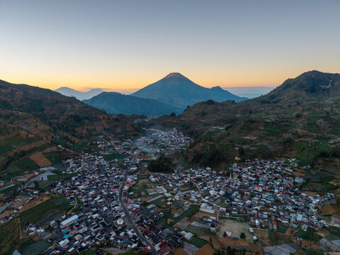 Aerial view of the village nestled between Mount Sindoro and Mount Sumbing with terraced agricultural fields under a soft golden hour sky in Kecamatan Kedu, Jawa Tengah, Indonesia.
