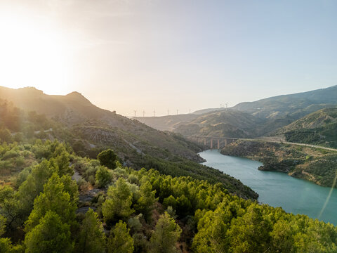 Aerial view of a reservoir surrounded by pine forests and rolling hills with a bridge and wind turbines in the distance, Velez de Benaudalla, Andalusia, Spain.
