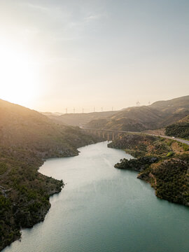 Aerial view of a winding river flowing through hills with a bridge and wind turbines under a golden sunset in Velez de Benaudalla, Andalusia, Spain.