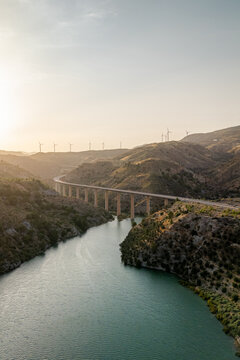 Aerial view of a modern bridge spanning a river in a hilly landscape with wind turbines on the horizon at sunset in Velez de Benaudalla, Andalusia, Spain.