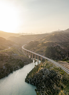 Aerial view of a highway bridge spanning a river in a hilly landscape with wind turbines on the horizon during golden hour in Velez de Benaudalla, Andalusia, Spain.