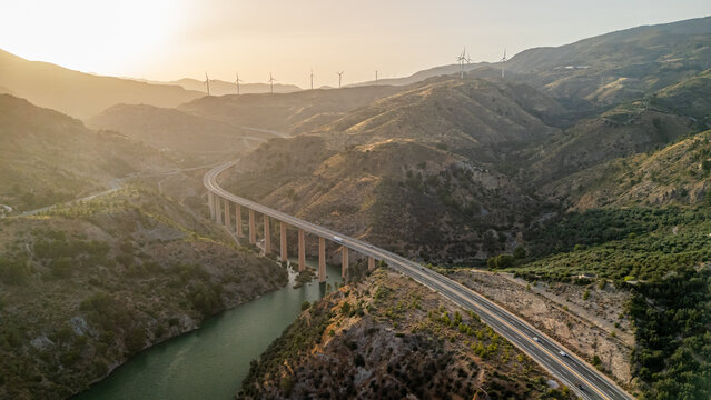 Aerial view of a highway bridge spanning a river in a mountainous landscape with wind turbines on the hills during sunset in Velez de Benaudalla, Andalusia, Spain.