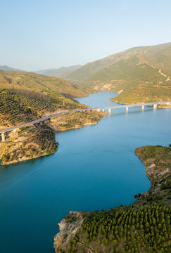 Aerial view of a bridge spanning a reservoir surrounded by rolling green hills and olive groves in Velez de Benaudalla, Andalusia, Spain.