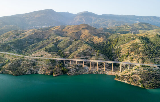 Aerial view of a modern viaduct bridge spanning across a turquoise reservoir, surrounded by rolling hills, olive groves, and rugged terrain in Velez de Benaudalla, Andalusia, Spain.