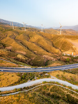 Aerial view of wind turbines on rolling golden hills with a highway and winding road in Velez de Benaudalla, Andalusia, Spain.