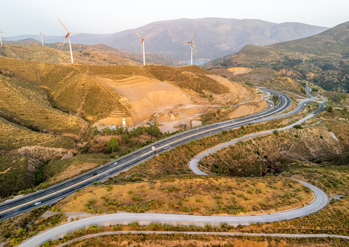 Aerial view of a highway winding through the arid, hilly landscape of Andalusia, featuring wind turbines on the ridges and sparse vegetation in Velez de Benaudalla, Andalusia, Spain.