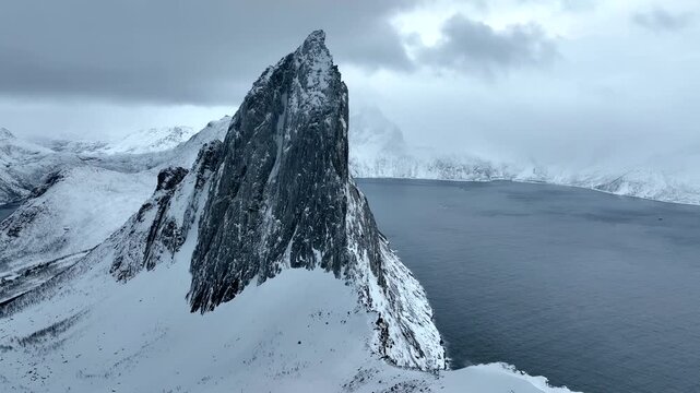 Aerial view of the dramatic snow-covered Segla mountain peak rising above the dark fjord waters under a cloudy sky in Senja, Troms, Norway.