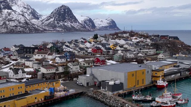 Aerial view of the colorful fishing village of Husoy with its harbor, fishing boats, and surrounding snow-capped mountains in Husoy, Troms, Norway.